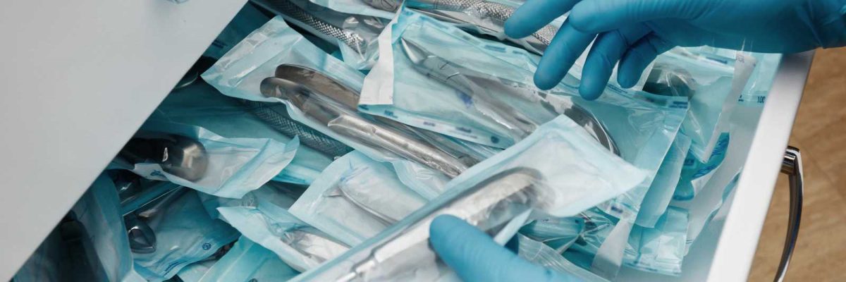 A close-up image of a person’s hands wearing blue gloves, sorting through various medical instruments which are packaged in individual sterile pouches. The instruments are neatly organized inside an open drawer, suggesting a clinic’s preparation for procedures.