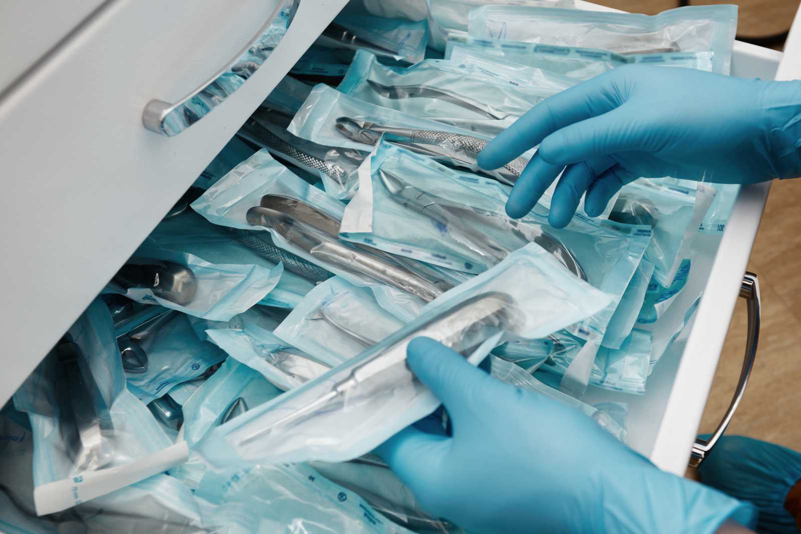 A close-up image of a person’s hands wearing blue gloves, sorting through various medical instruments which are packaged in individual sterile pouches. The instruments are neatly organized inside an open drawer, suggesting a clinic’s preparation for procedures.